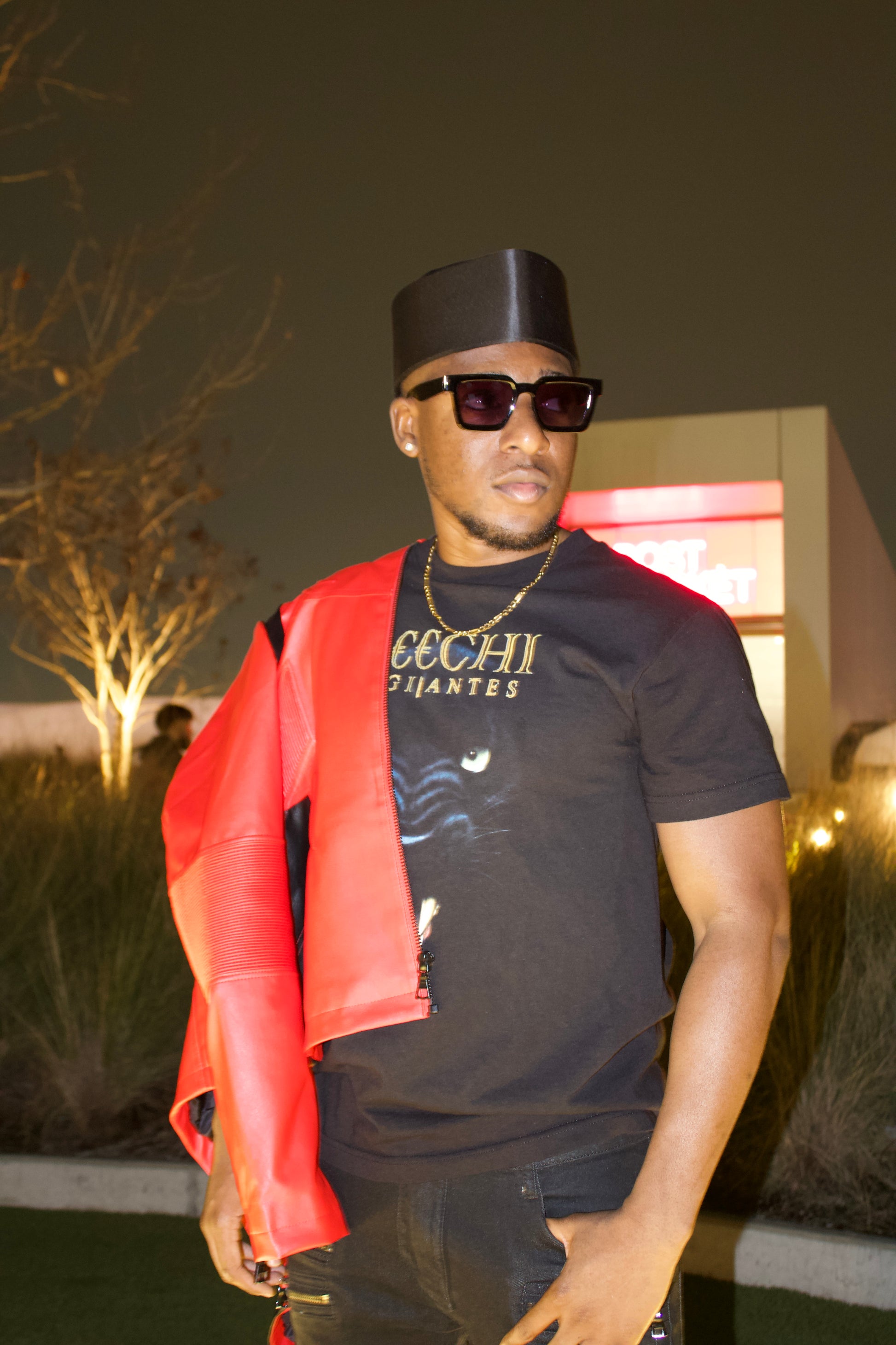Man wearing an African native black cap, sunglasses, and a black t-shirt with red jacket outdoors at night.