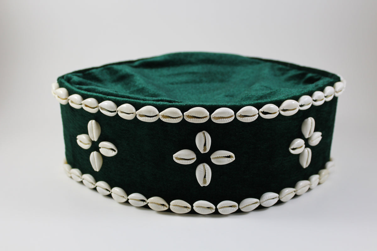A dark green velvet cylindrical hat or cap decorated with white cowrie shells along the edges and in floral patterns on the front, set against a plain light gray background.