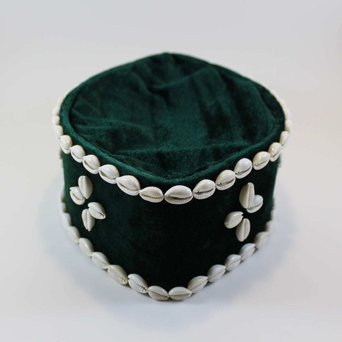 A dark green velvet cylindrical hat or pouch decorated with white cowrie shells around the rim and on the front, set against a plain light background.