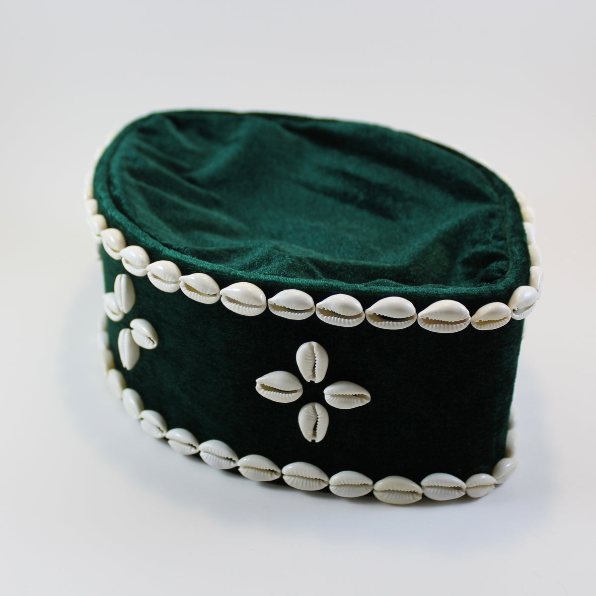 A dark green velvet cap decorated with white cowrie shells around the border and a central cowrie shell flower motif on the front, set against a plain light background.